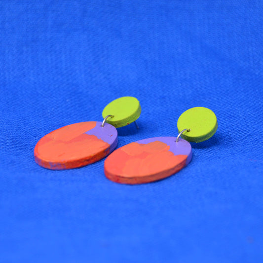 A pair of handmade wooden dangle earrings with abstract painted designs in orange, purple, and green, displayed on a textured blue background.