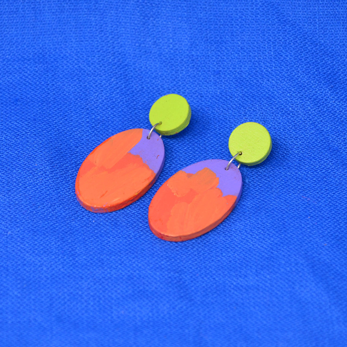 A pair of handmade drop earrings with lime green circular studs and oval dangles painted in orange and purple brushstroke patterns, displayed on a textured blue background.