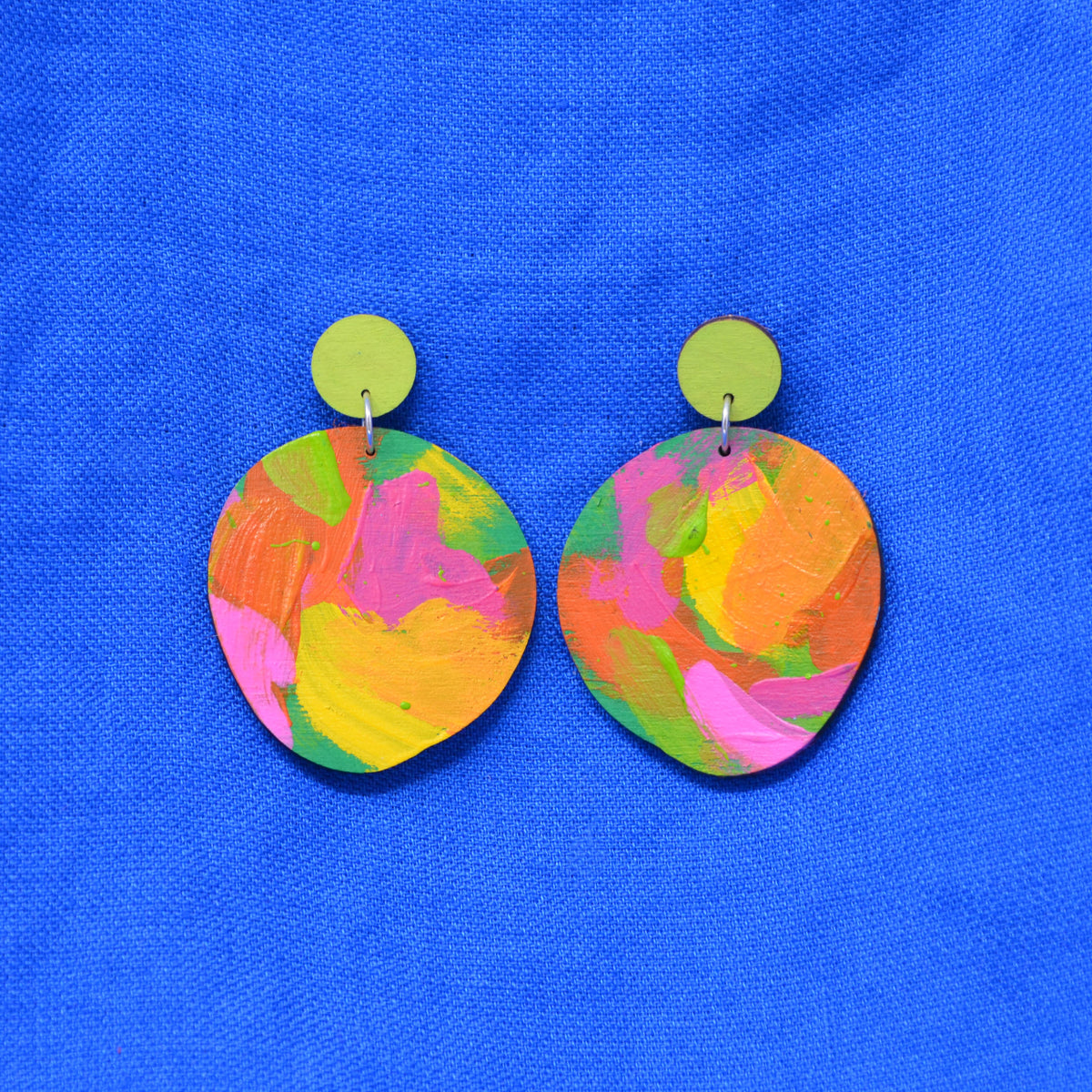 A pair of handmade drop earrings with irregular, hand-painted wooden discs in a vibrant multicolor pattern of pink, yellow, green, and orange, hanging from lime green circular studs against a bright blue textured background.