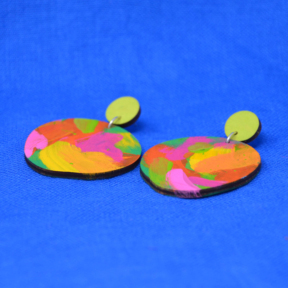 A pair of handmade drop earrings with irregular, hand-painted wooden discs in a vibrant multicolor pattern of pink, yellow, green, and orange, hanging from lime green circular studs against a bright blue textured background.