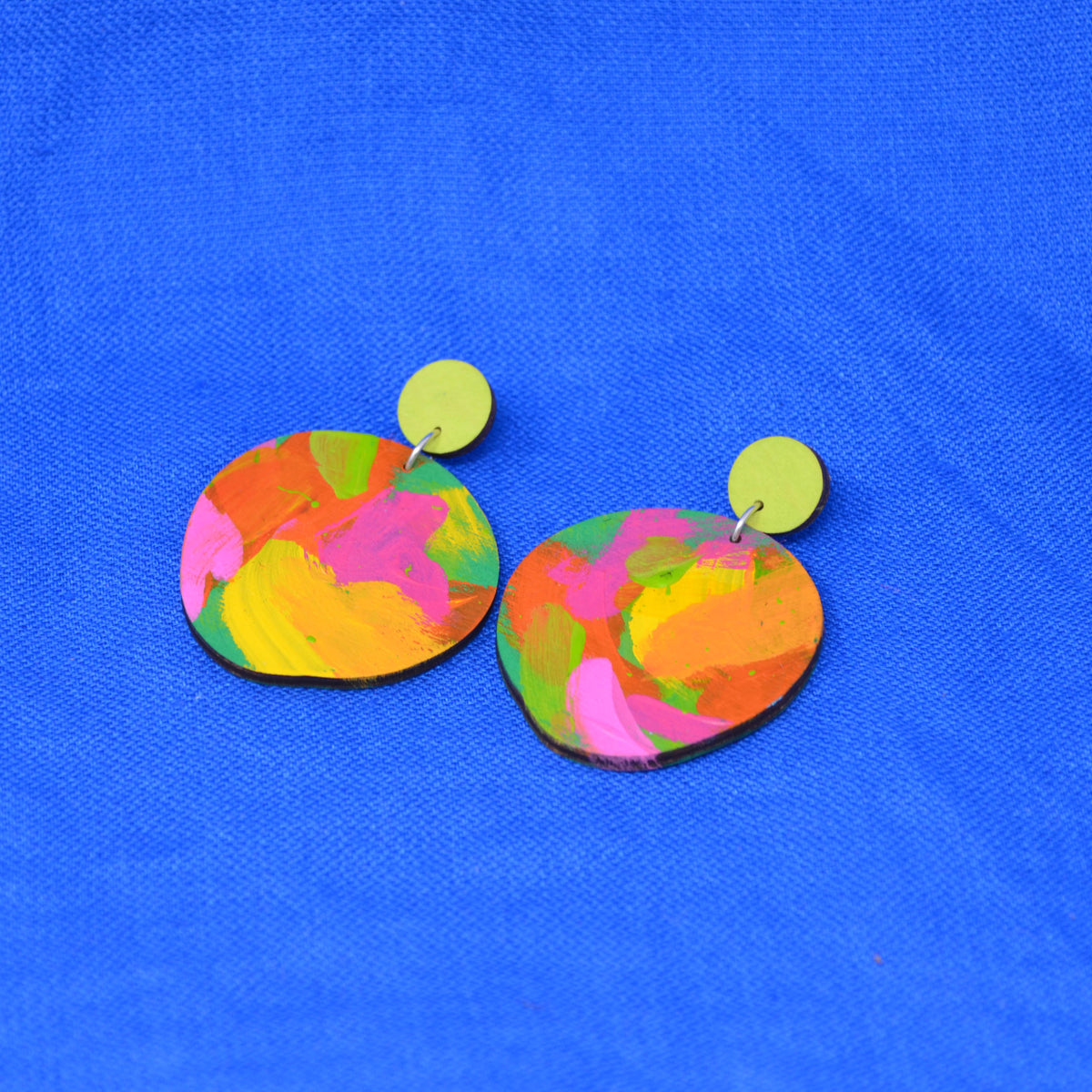 A pair of handmade drop earrings with irregular, hand-painted wooden discs in a vibrant multicolor pattern of pink, yellow, green, and orange, hanging from lime green circular studs against a bright blue textured background.