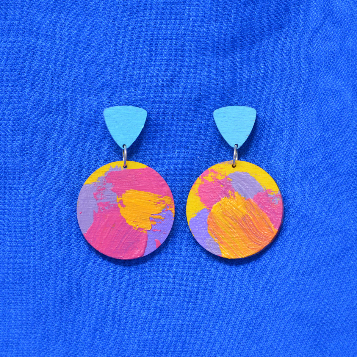 A pair of drop earrings with light blue triangular studs and circular dangles painted with abstract brushstrokes of pink, yellow, and purple, displayed on a textured blue background.