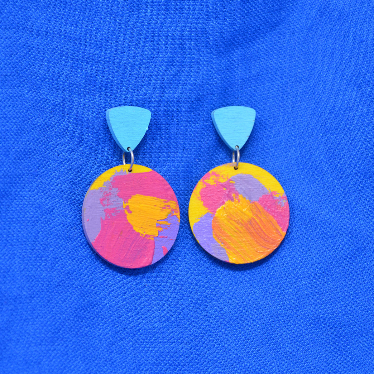 A pair of drop earrings with light blue triangular studs and circular wooden pendants featuring an abstract multicolor paint design in pink, yellow, orange, and purple, displayed on a textured blue fabric background.