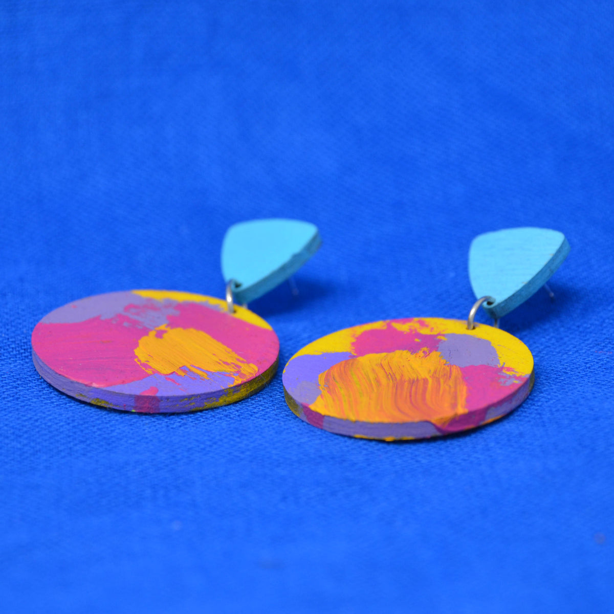 A pair of drop earrings with light blue triangular-shaped studs and round disc charms featuring an abstract multicolor paint design in pink, yellow, and orange, set against a solid blue background.