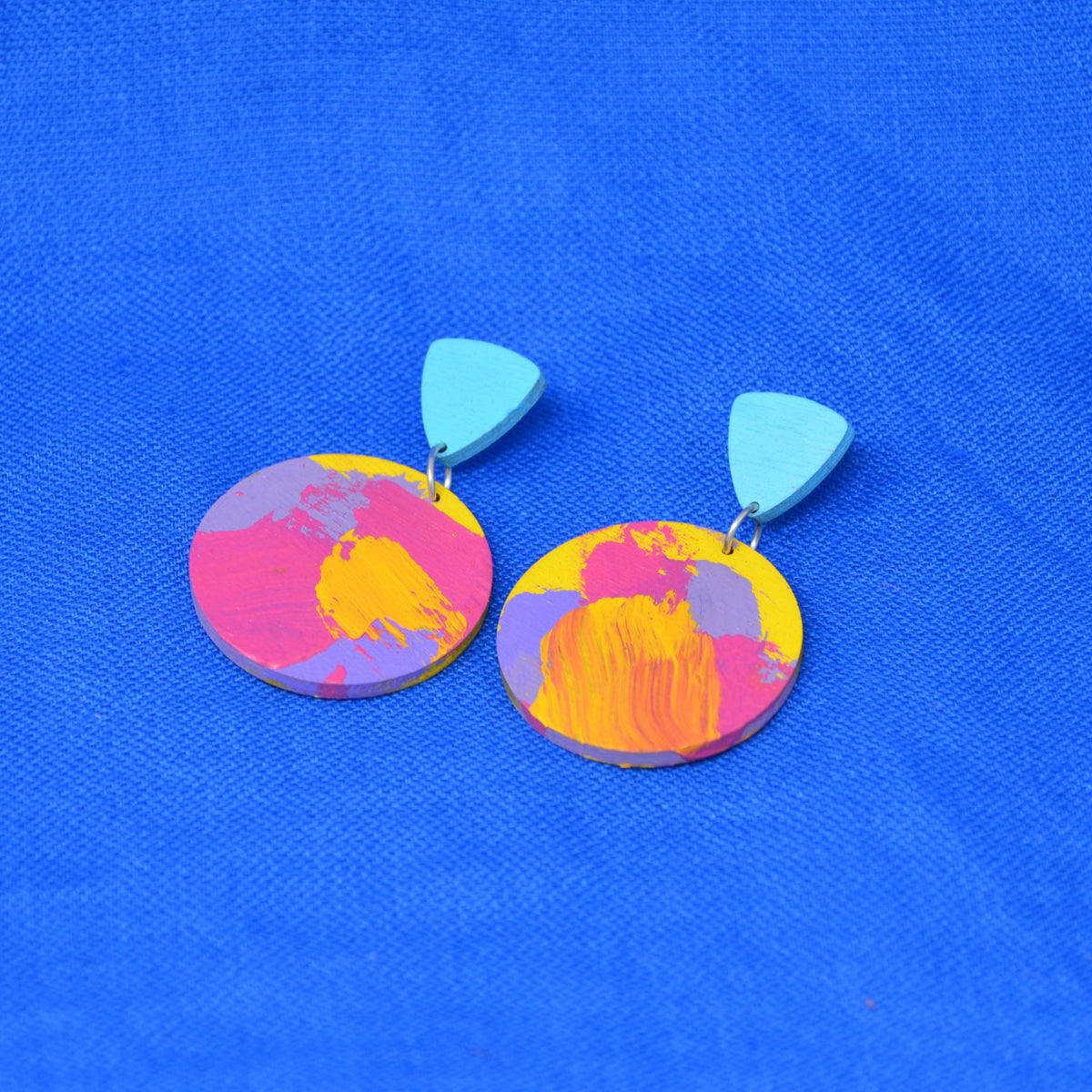 A pair of drop earrings with light blue triangular top pieces and round wooden disc charms featuring a colorful abstract paint design in pink, yellow, and purple, displayed on a textured blue background.