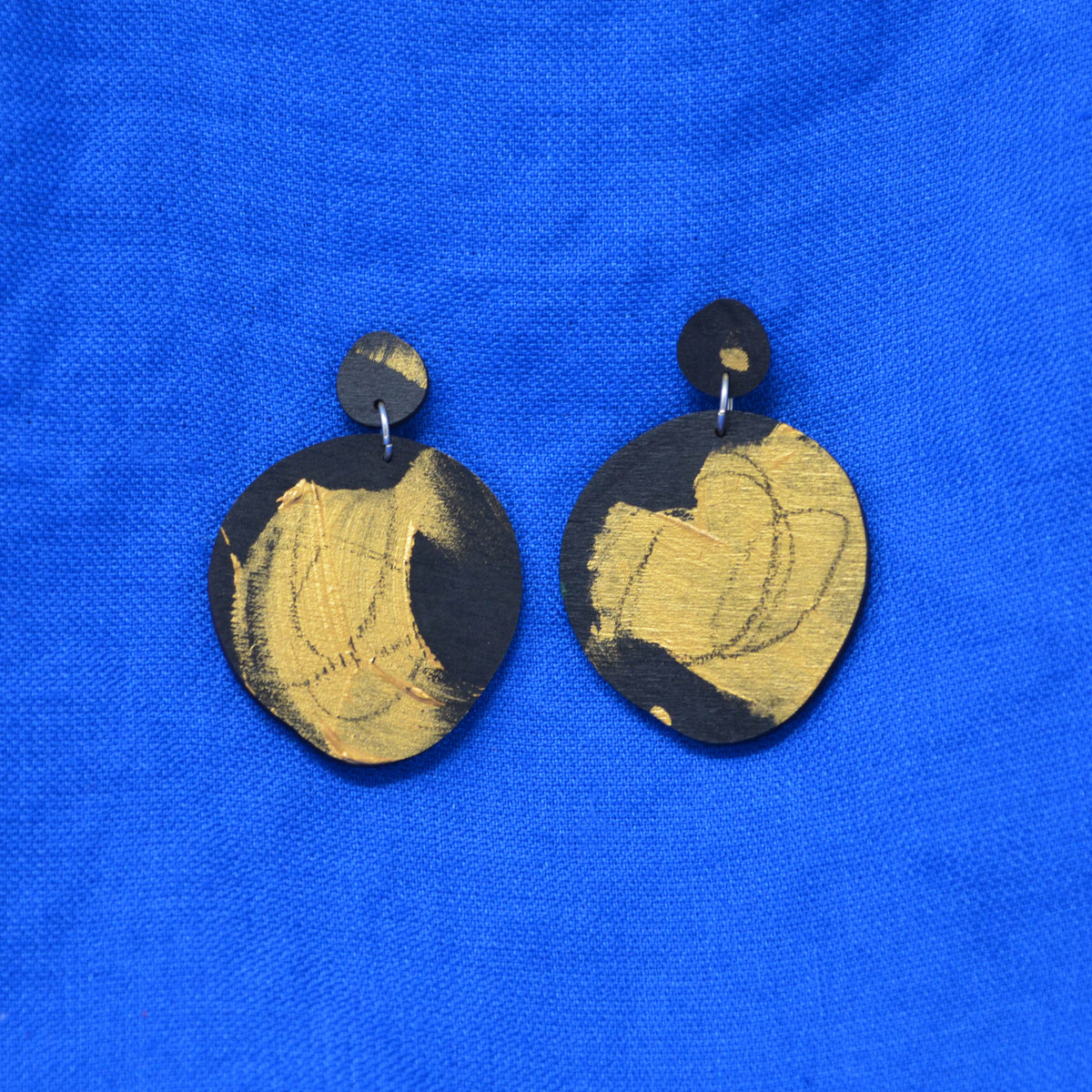 A pair of handmade wooden dangle earrings with a black and gold abstract painted design, displayed on a vibrant blue textured fabric background.