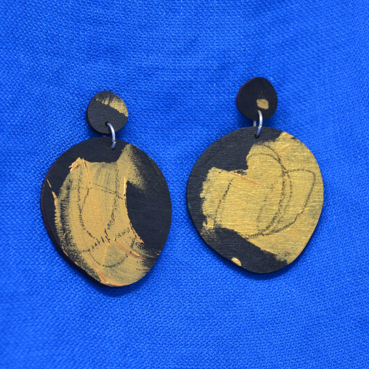 A pair of hand-painted wooden earrings with a black base and gold  accents, displayed on a vibrant blue textured background.