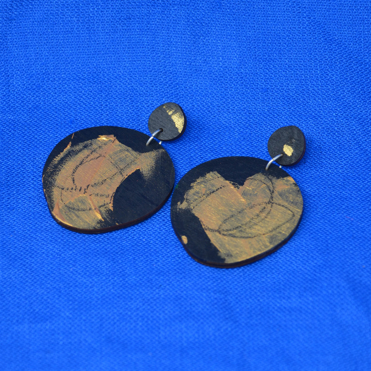 A pair of handmade dangle earrings made from irregular black discs with gold patterns, shown on a textured blue background.