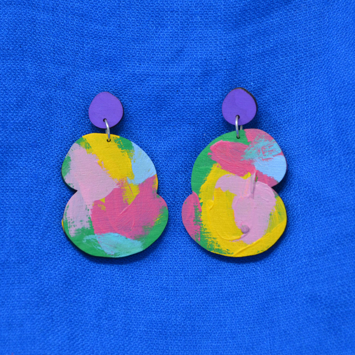 A pair of abstract drop earrings with a vibrant pink, yellow, blue and green abstract paint design, attached to purple fronted stud, displayed on a textured blue background.