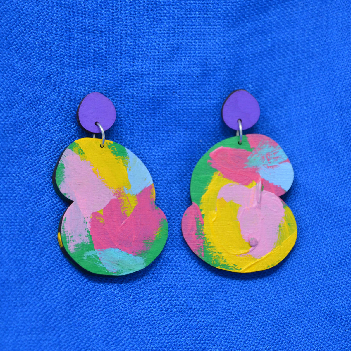 A pair of abstract drop earrings with a vibrant pink, yellow, blue and green abstract paint design, attached to purple fronted stud, displayed on a textured blue background.