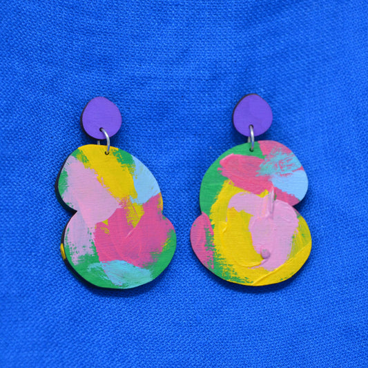 A pair of abstract drop earrings with a vibrant pink, yellow, blue and green abstract paint design, attached to purple fronted stud, displayed on a textured blue background.