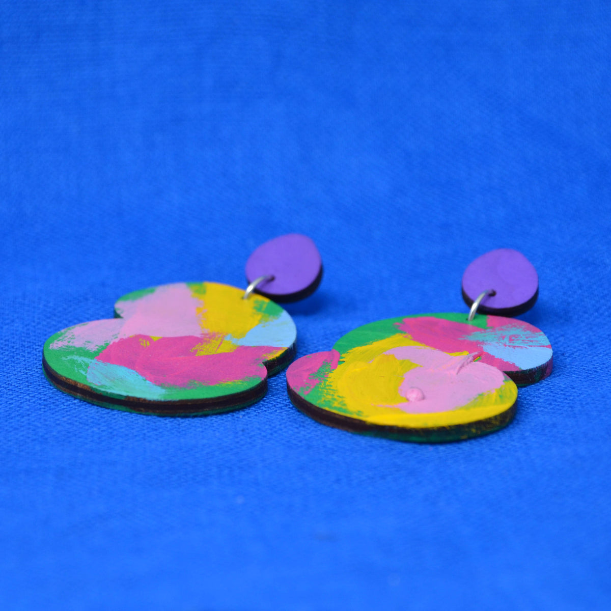 A pair of abstract drop earrings with a vibrant pink, yellow, blue and green abstract paint design, attached to purple fronted stud, displayed on a textured blue background.