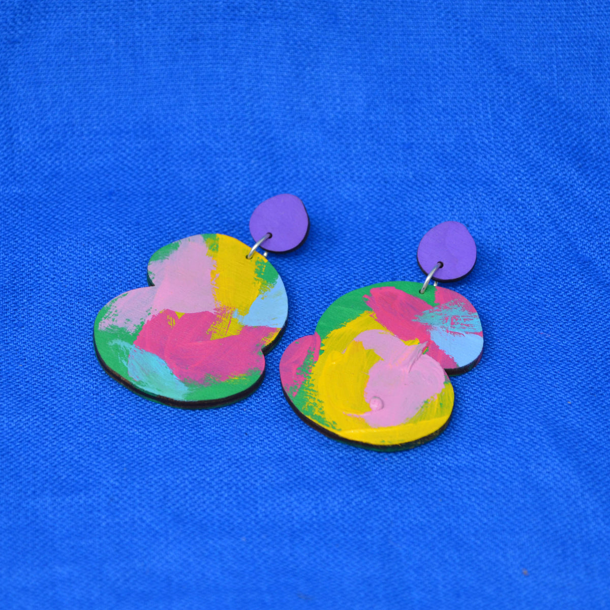A pair of abstract drop earrings with a vibrant pink, yellow, blue and green abstract paint design, attached to purple fronted stud, displayed on a textured blue background.