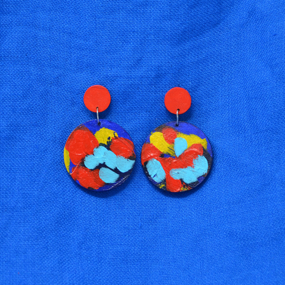 A pair of handmade drop earrings with bright red circular studs and circle dangles featuring a blue, red and yellow abstract painted design, displayed on a textured blue background.