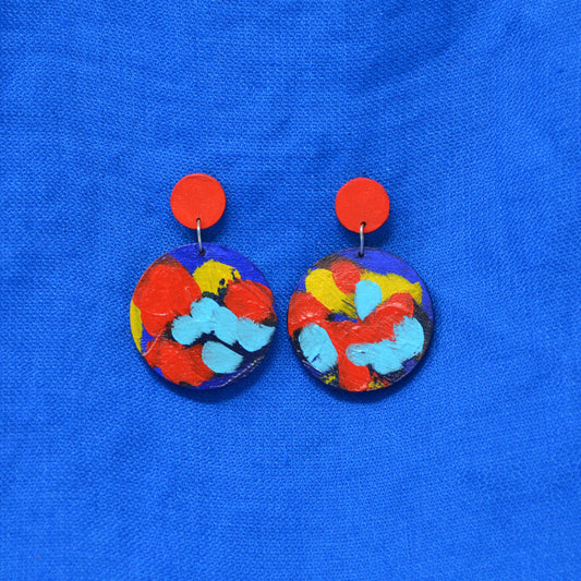 A pair of handmade drop earrings with bright red circular studs and circle dangles featuring a blue, red and yellow abstract painted design, displayed on a textured blue background.