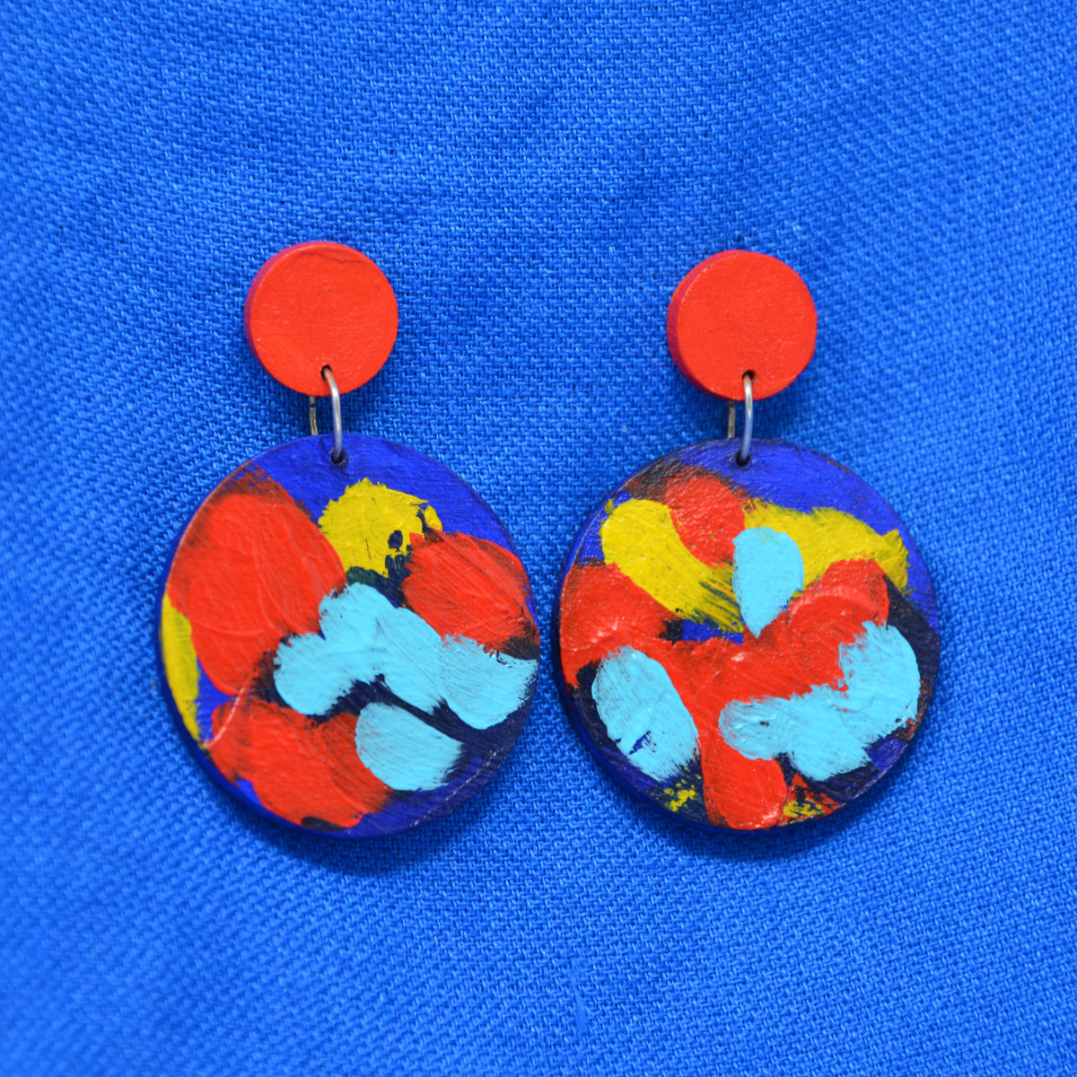 A pair of handmade drop earrings with bright red circular studs and circle dangles featuring a blue, red and yellow abstract painted design, displayed on a textured blue background.