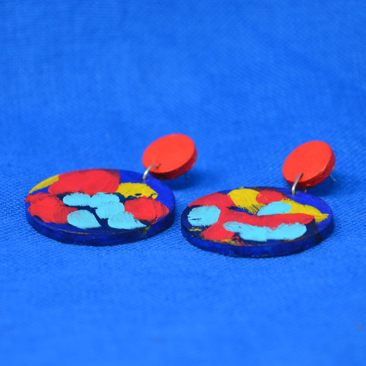 A pair of handmade drop earrings with bright red circular studs and circle dangles featuring a blue, red and yellow abstract painted design, displayed on a textured blue background.