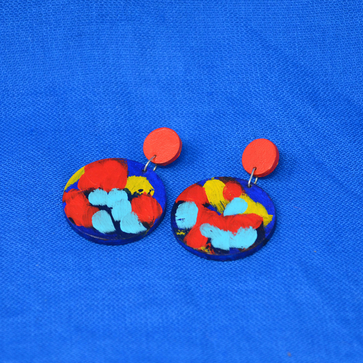 A pair of handmade drop earrings with bright red circular studs and circle dangles featuring a blue, red and yellow abstract painted design, displayed on a textured blue background.