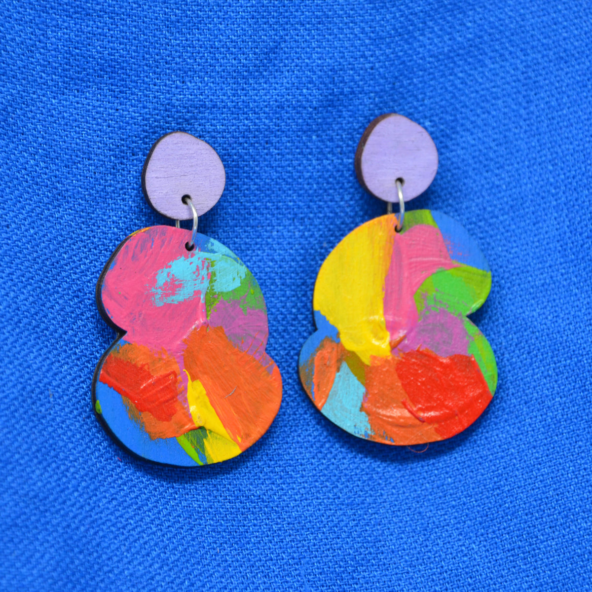 A pair of colorful abstract acrylic earrings with a light purple circular stud and a larger multicolor painted drop piece, displayed on a textured blue background.