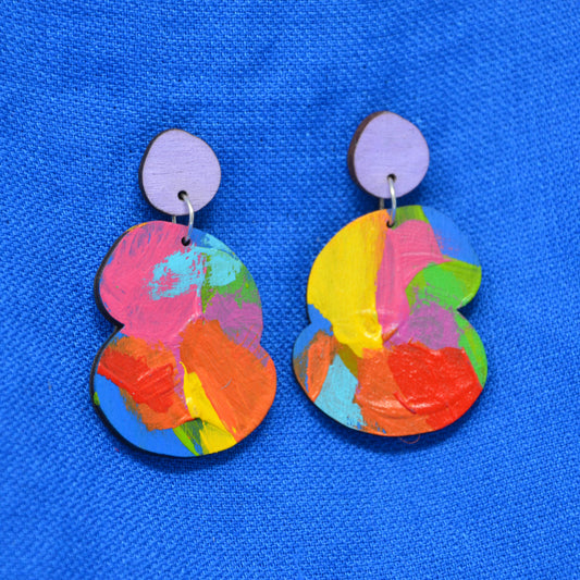 A pair of colorful abstract acrylic earrings with a light purple circular stud and a larger multicolor painted drop piece, displayed on a textured blue background.