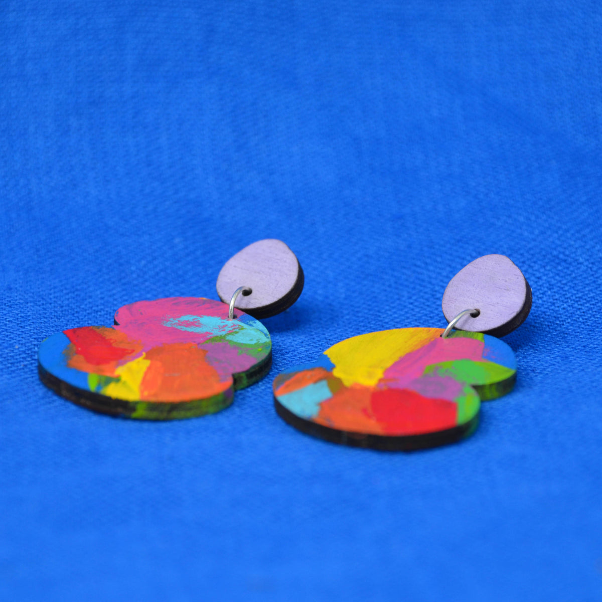 A pair of colorful wooden dangle earrings with abstract painted patterns in pink, yellow, red, green, and blue, featuring light purple oval studs, displayed on a textured blue background.