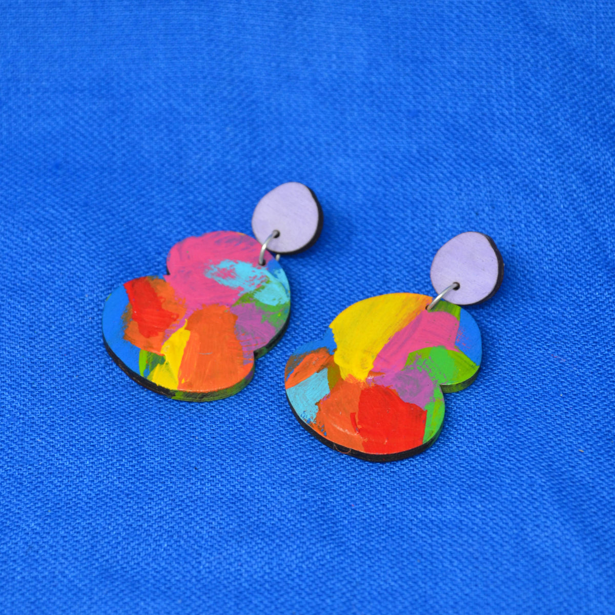 A pair of colorful abstract acrylic drop earrings with light purple circular studs, displayed on a textured blue background.