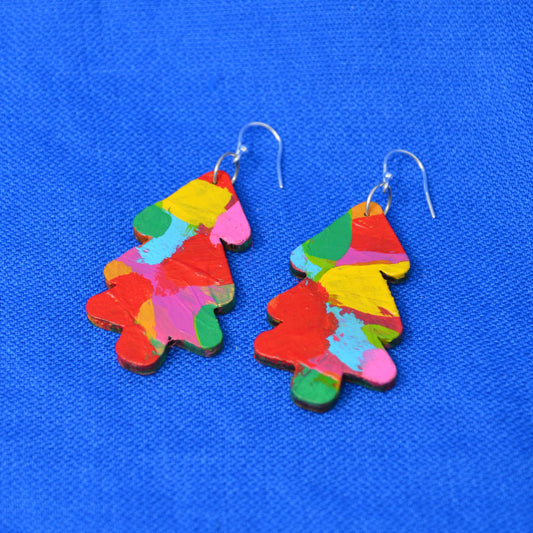 A pair of tree-shaped drop earrings painted with a multicolor abstract brushstroke pattern, hanging from silver fishhook wires on a textured blue background.