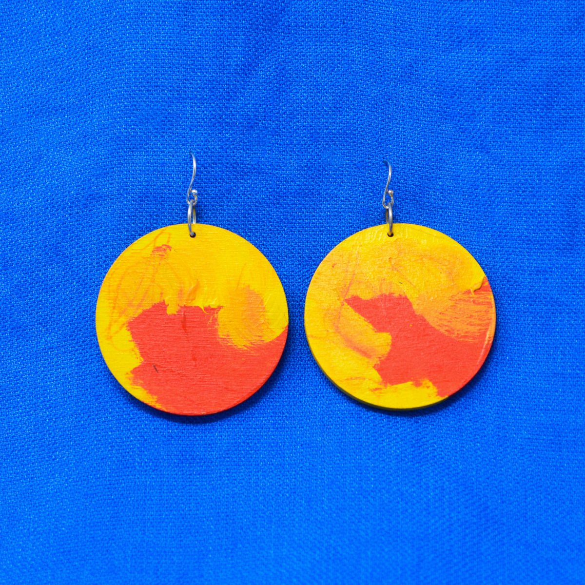 A pair of round wooden drop earrings painted with yellow and orange abstract brushstrokes, hanging from silver fishhook wires against a bright blue textured fabric background.