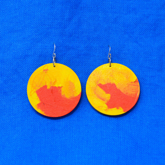A pair of round wooden drop earrings painted with yellow and orange abstract brushstrokes, hanging from silver fishhook wires against a bright blue textured fabric background.