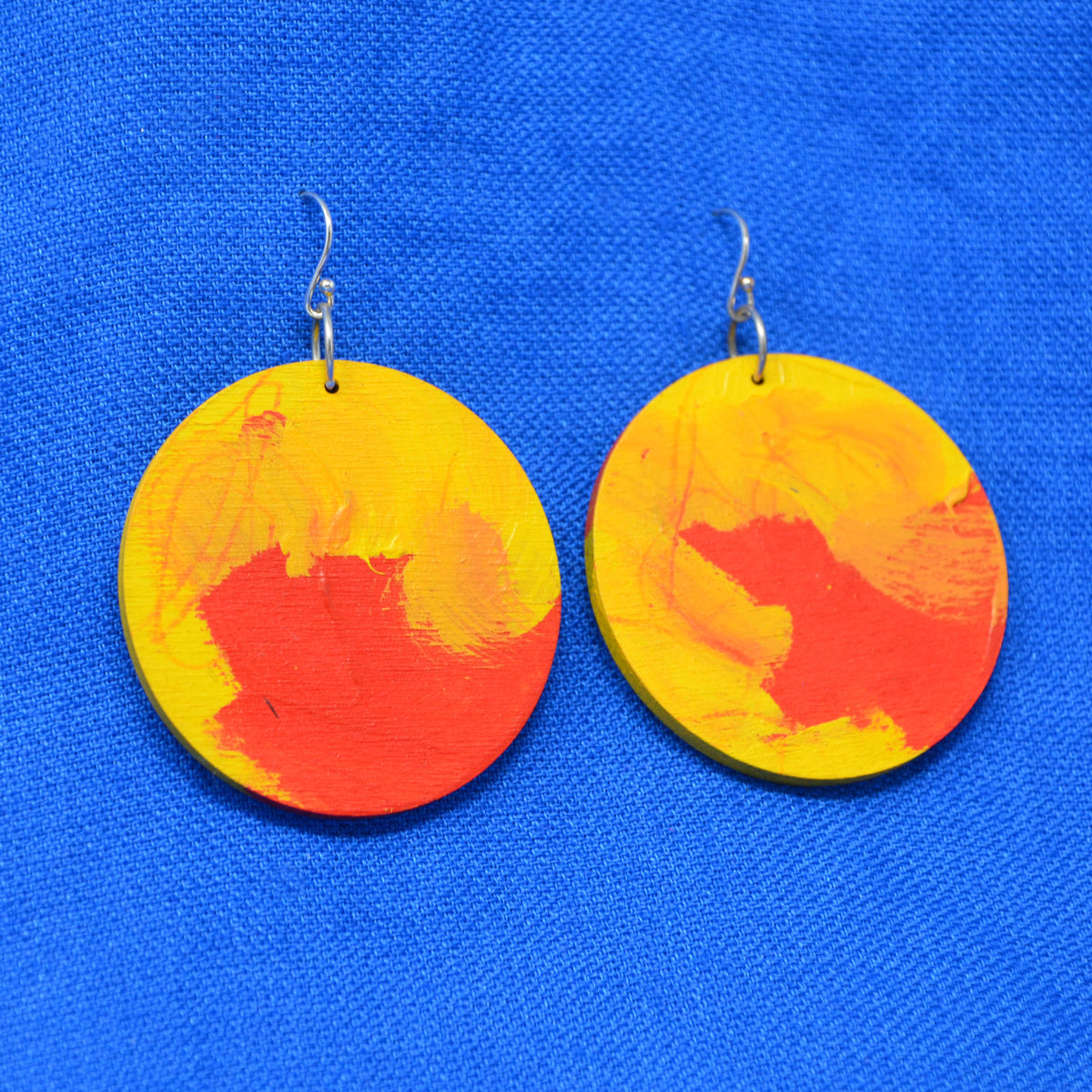 A pair of round wooden dangle earrings painted with yellow and orange-red abstract brushstrokes, hanging from silver fishhook wires against a bright blue textured fabric background.