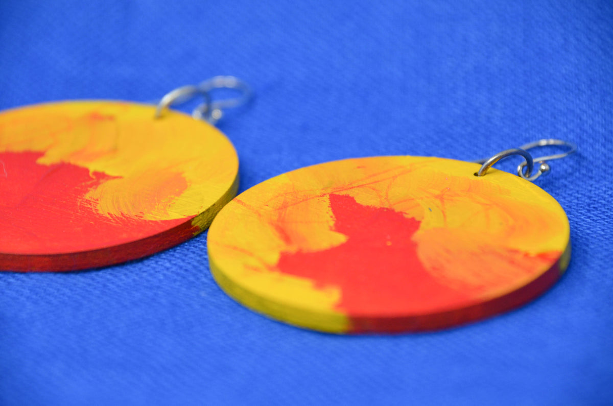 A pair of round wooden drop earrings painted with a red and yellow abstract design, hanging from silver hooks against a textured blue background.