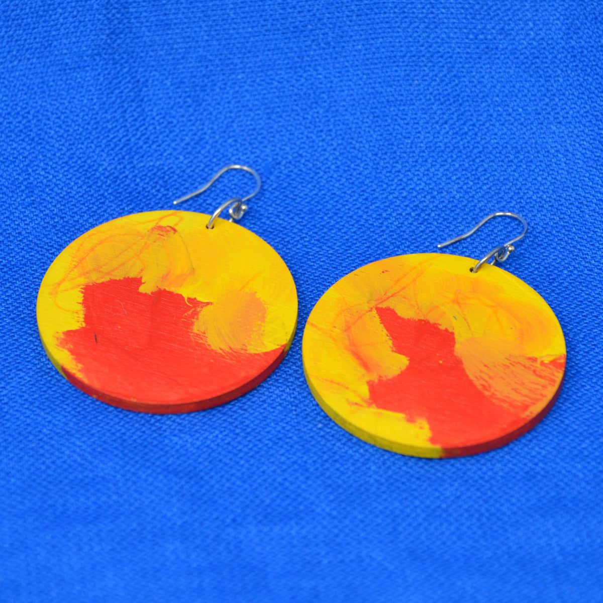 A pair of round wooden drop earrings painted with yellow and red brushstrokes, hanging from silver fishhook wires on a bright blue textured fabric background.
