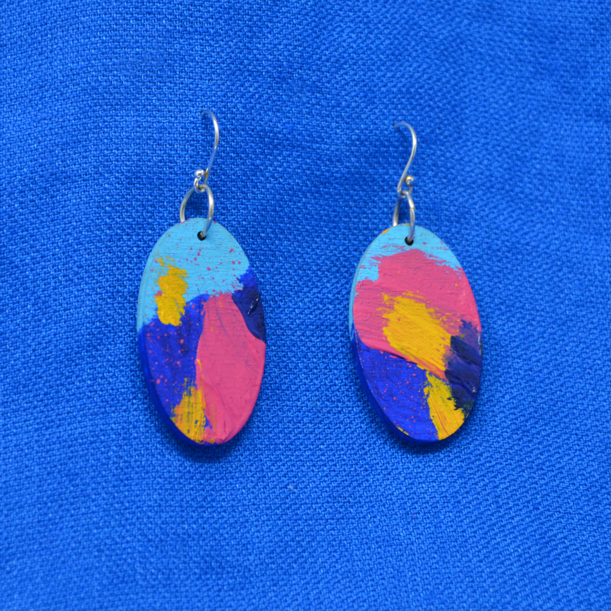 A pair of oval drop earrings with a colorful abstract paint design in pink, blue, yellow, and light blue, hanging from silver fishhook wires on a textured blue background.