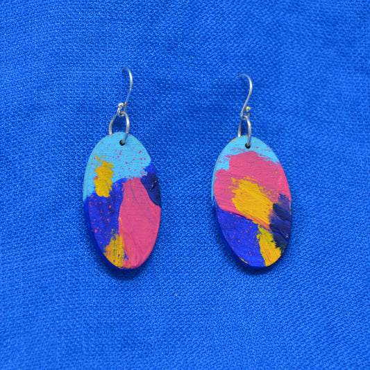 A pair of oval drop earrings with a colorful abstract paint design in pink, blue, yellow, and light blue, hanging from silver fishhook wires on a textured blue background.