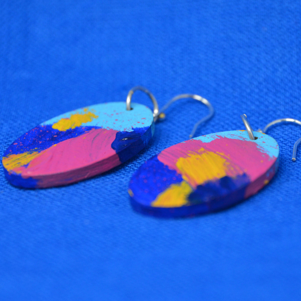 A pair of oval drop earrings with a colorful abstract paint design in pink, blue, yellow, and light blue, hanging from silver fishhook wires on a textured blue background.