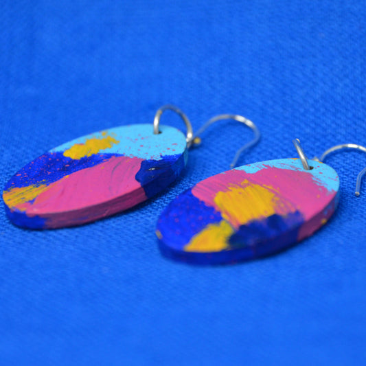 A pair of oval drop earrings with a colorful abstract paint design in pink, blue, yellow, and light blue, hanging from silver fishhook wires on a textured blue background.