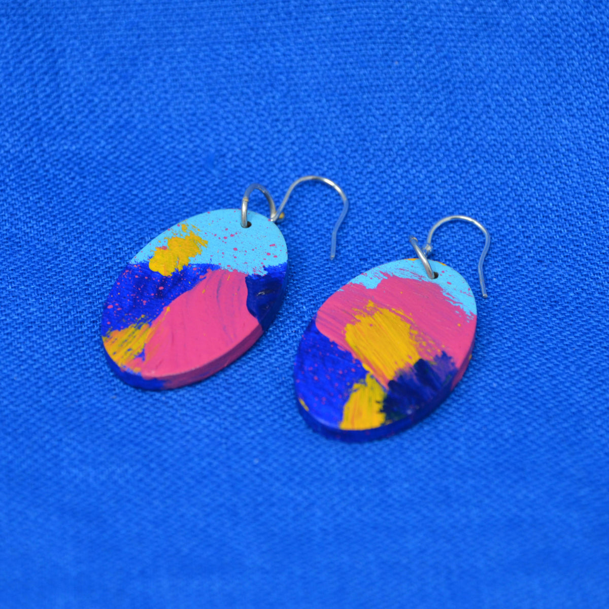 A pair of oval drop earrings with a colorful abstract paint design in pink, blue, yellow, and light blue, hanging from silver fishhook wires on a textured blue background.
