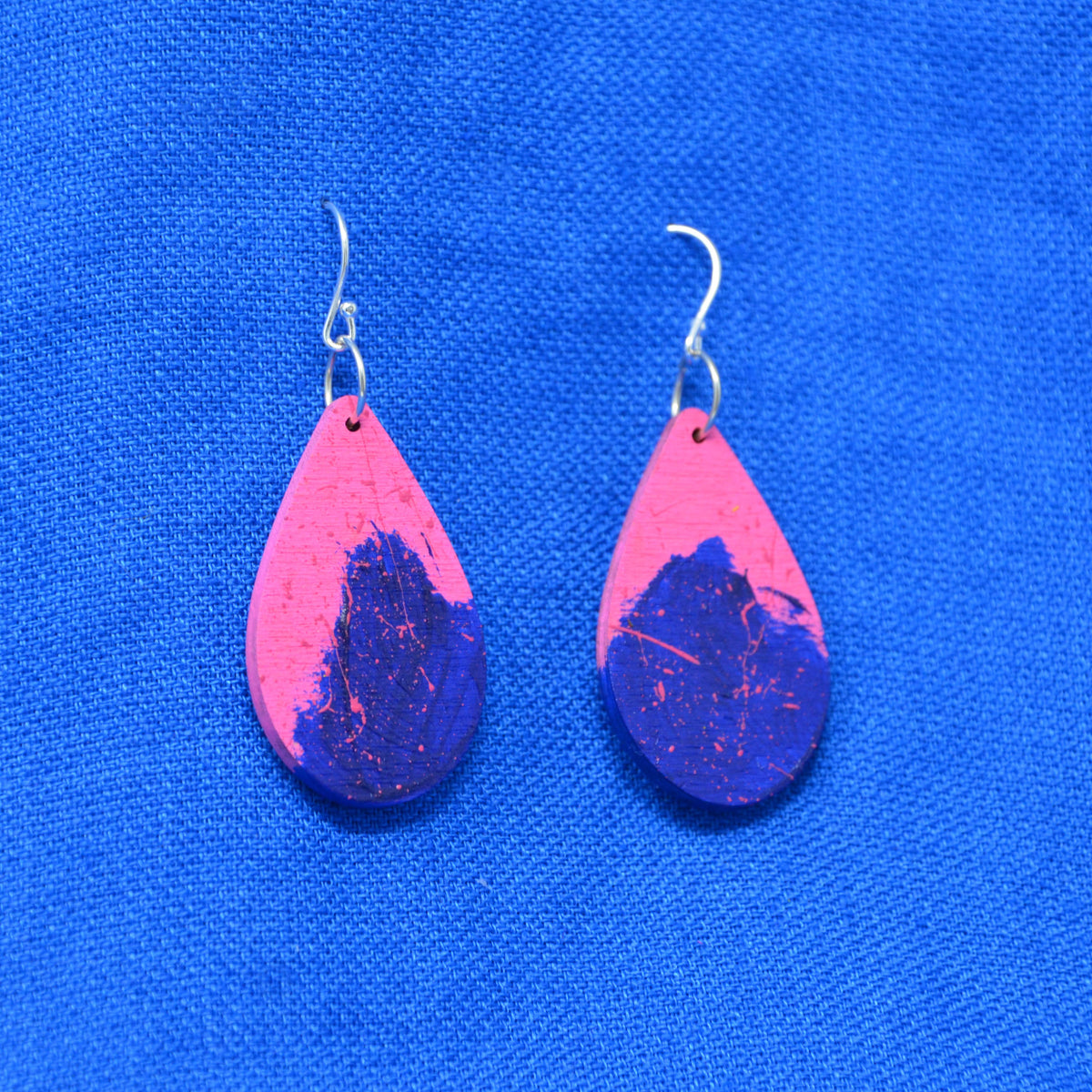 A pair of teardrop-shaped dangle earrings with a bright pink and deep blue marbled pattern, suspended from silver fishhook wires, displayed on a textured blue background.