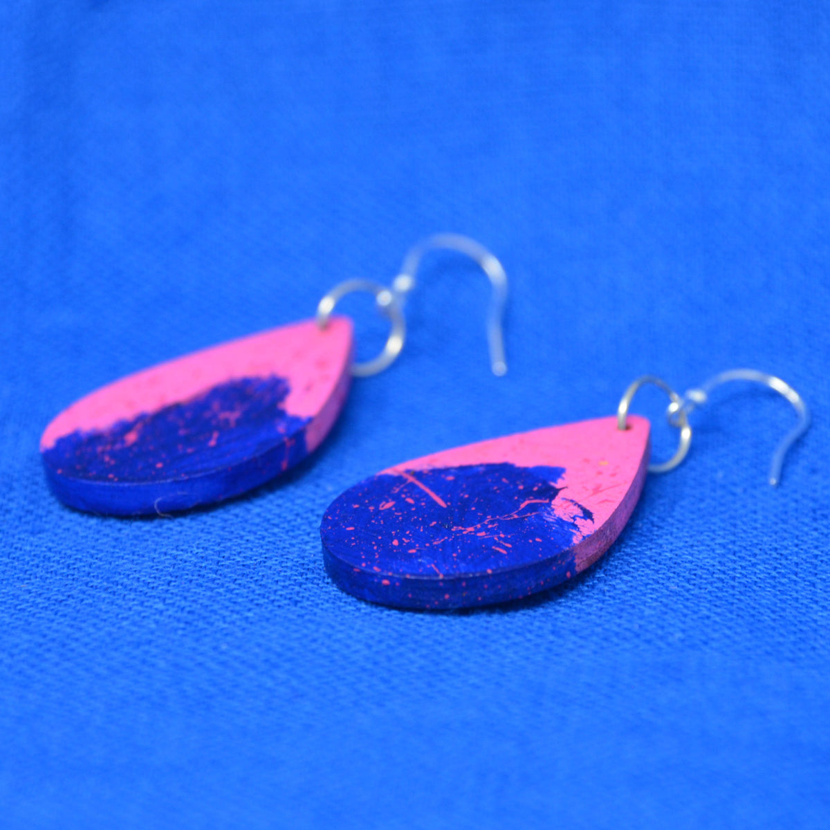 A pair of teardrop-shaped wooden earrings with a split pink and blue design, featuring silver fishhook ear wires, displayed on a textured blue background.
