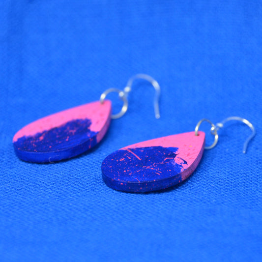 A pair of teardrop-shaped wooden earrings with a split pink and blue design, featuring silver fishhook ear wires, displayed on a textured blue background.