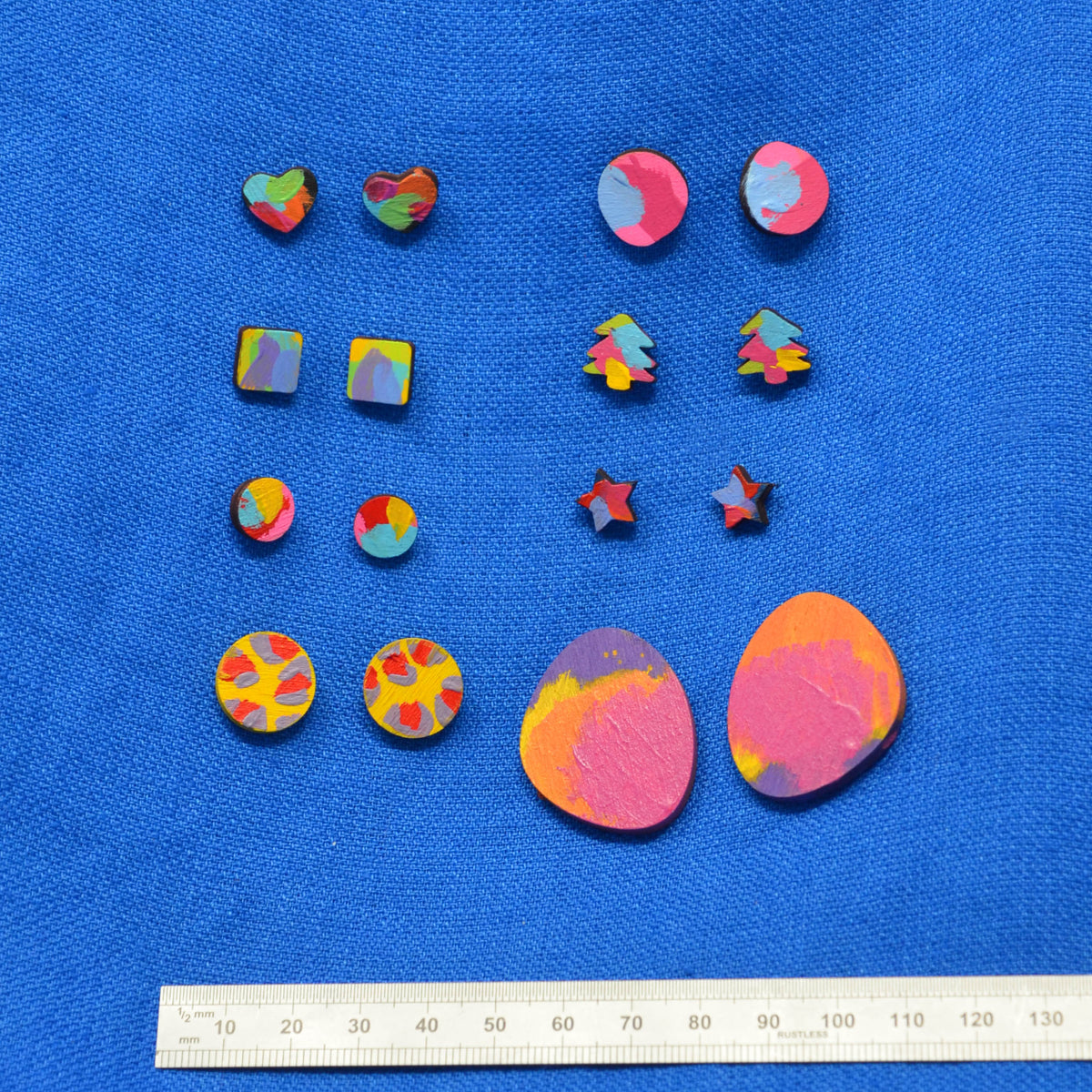 A set of colorful wooden stud earrings in various shapes (hearts, squares, stars, trees, and large teardrop pieces) displayed on a blue textured background with a ruler at the bottom for scale.