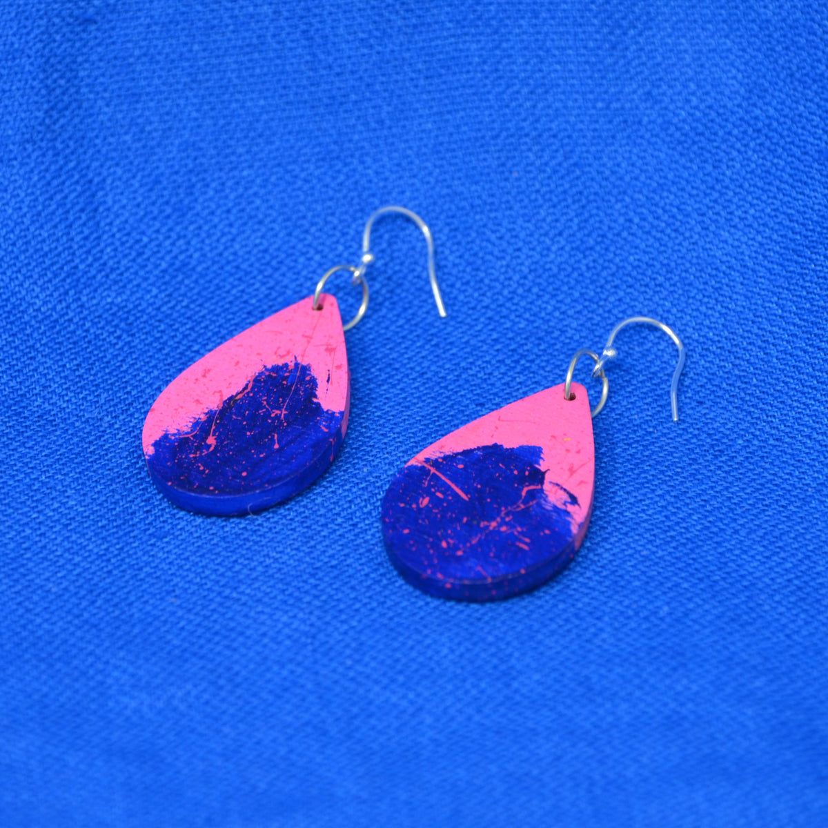 A pair of teardrop-shaped dangle earrings with a vibrant pink and deep blue split design, featuring pink flecks, displayed on a textured blue background.