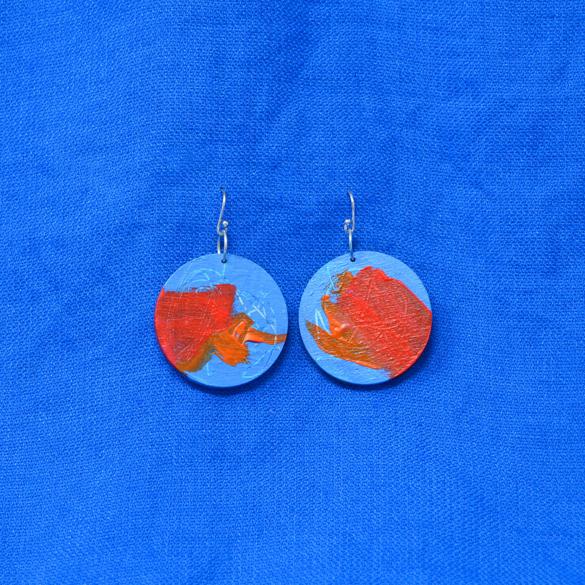 A pair of handmade round dangle earrings featuring a blue and orange abstract painted design, suspended from silver fishhook wires, displayed on a textured blue fabric background.