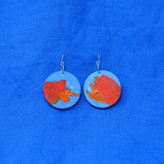 A pair of handmade round dangle earrings featuring a blue and orange abstract painted design, suspended from silver fishhook wires, displayed on a textured blue fabric background.