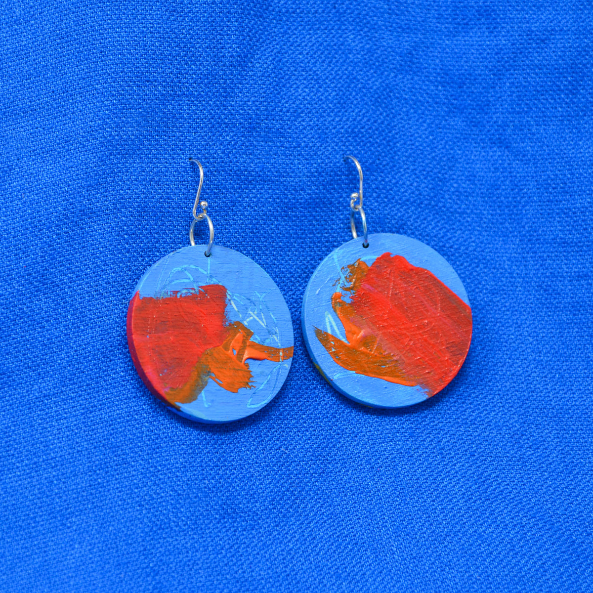A pair of round drop earrings with silver fishhook wires, featuring abstract painted designs in red, orange, and light blue, displayed on a textured blue fabric background.