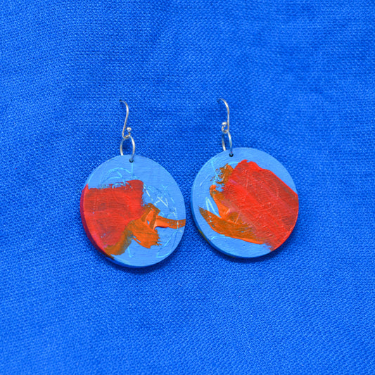 A pair of round drop earrings with silver fishhook wires, featuring abstract painted designs in red, orange, and light blue, displayed on a textured blue fabric background.