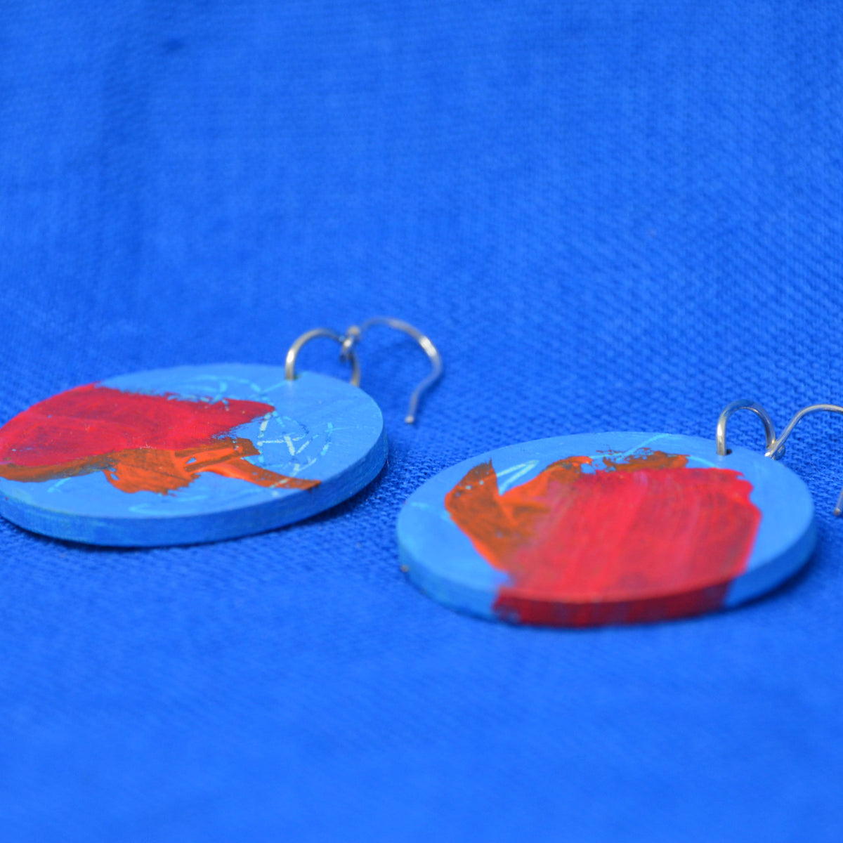 A pair of handmade round wooden earrings painted with bright blue and red brushstrokes, featuring silver fishhook ear wires, displayed on a textured blue background.