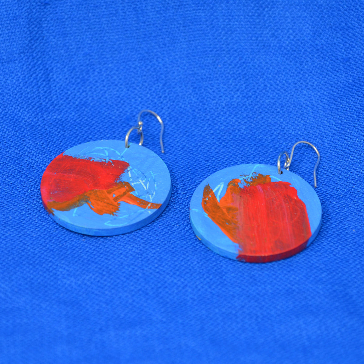 A pair of round blue wooden earrings with abstract red and orange painted brushstrokes, hanging from silver fishhook wires on a textured blue background.