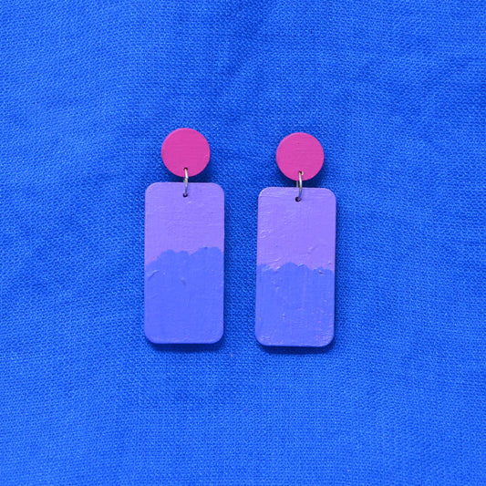 A pair of geometric drop earrings with bright pink circular studs and rectangular dangles in purple and blue, displayed on a textured blue background.