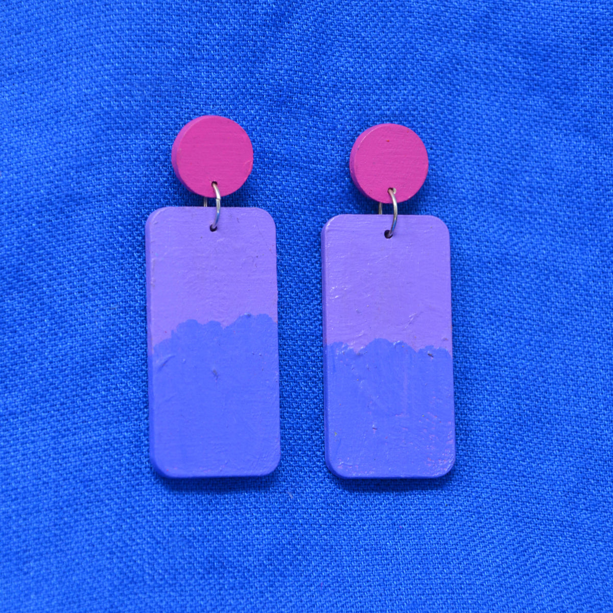 A pair of handmade drop earrings featuring rectangular purple and blue painted wooden charms with bright pink circular studs, displayed on a textured blue fabric background.